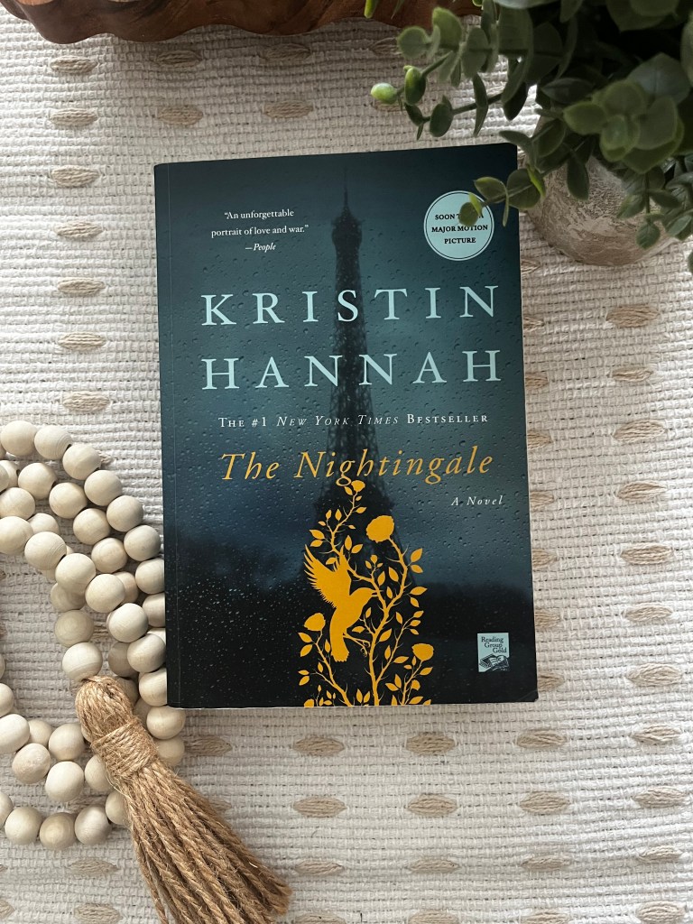 The Nightingale by Kristin Hannah displayed on a cream textured blanket with wooden beads and greenery.
