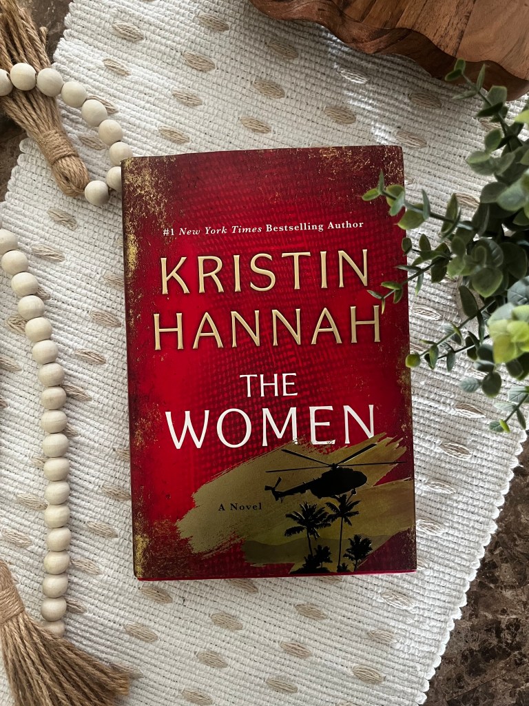 The Women by Kristin Hannah displayed on a cream textured fabric with wooden beads and greenery.