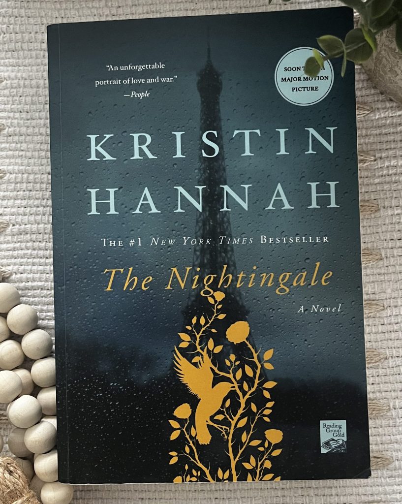The Nightingale by Kristin Hannah displayed on a cream textured blanket with wooden beads and greenery.