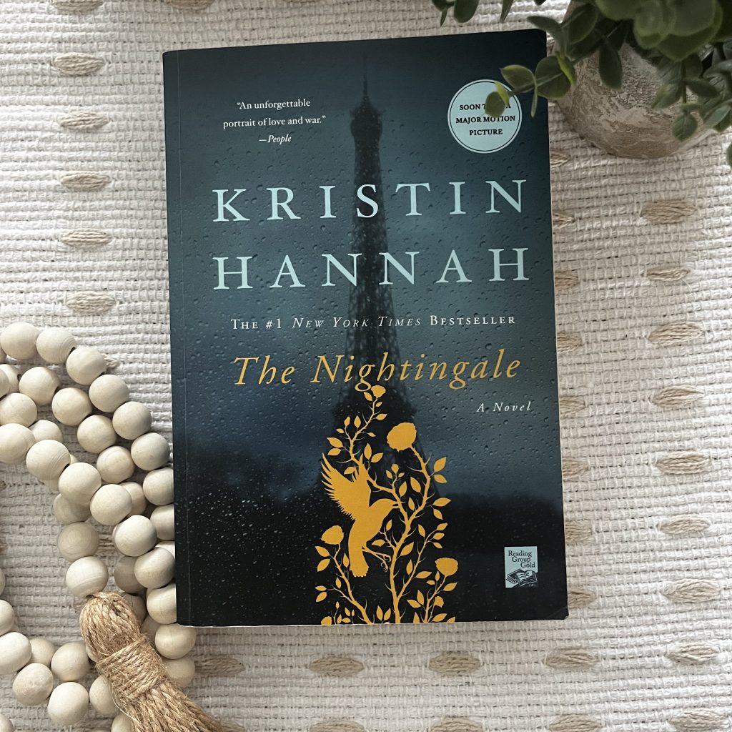 The Nightingale by Kristin Hannah displayed on a cream textured blanket with wooden beads and greenery.