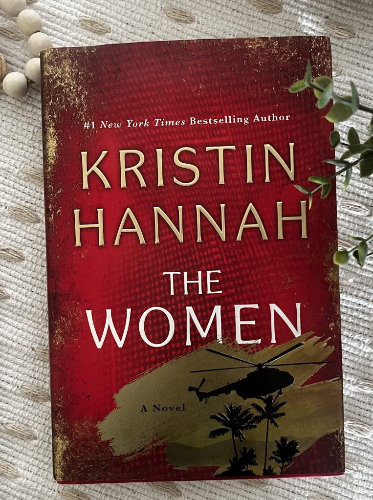 The Women by Kristin Hannah displayed on a cream textured fabric with wooden beads and greenery.