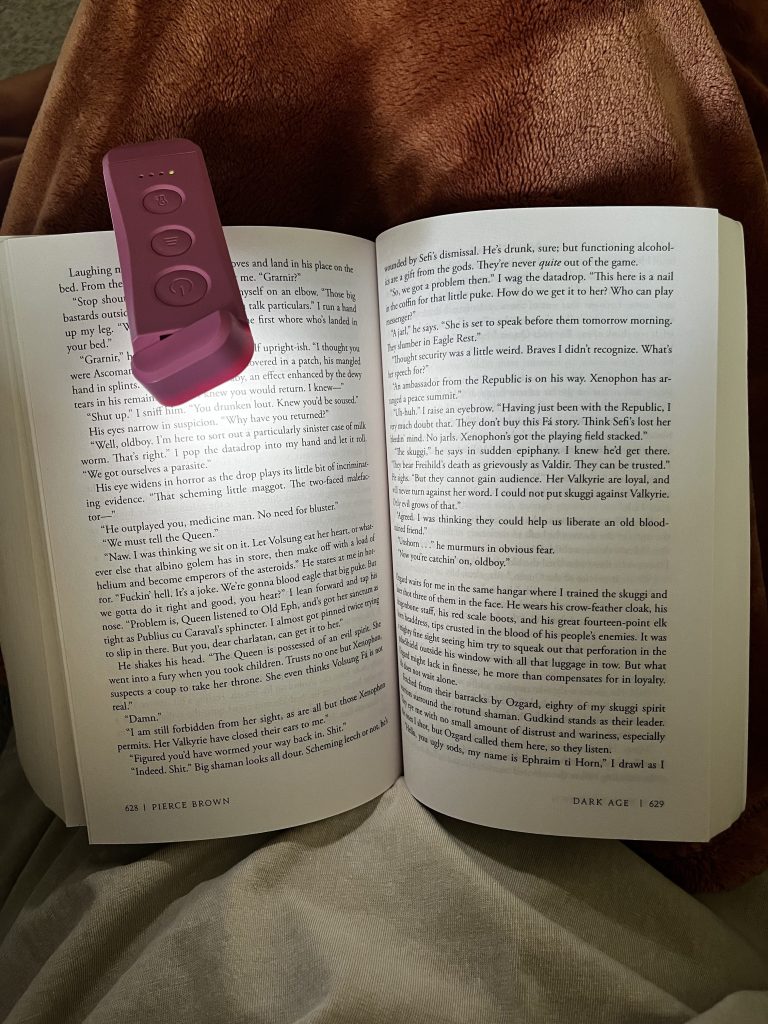 Pink clip-on book light attached to an open book while reading under a blanket.