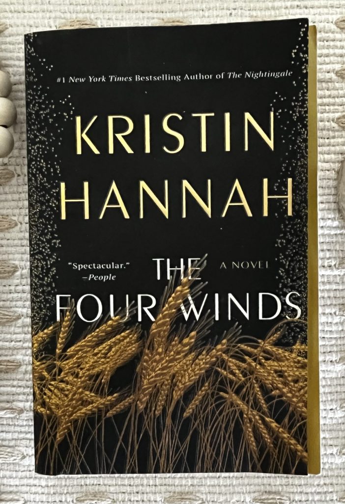 The Four Winds by Kristin Hannah on a cream textured blanket with wooden beads