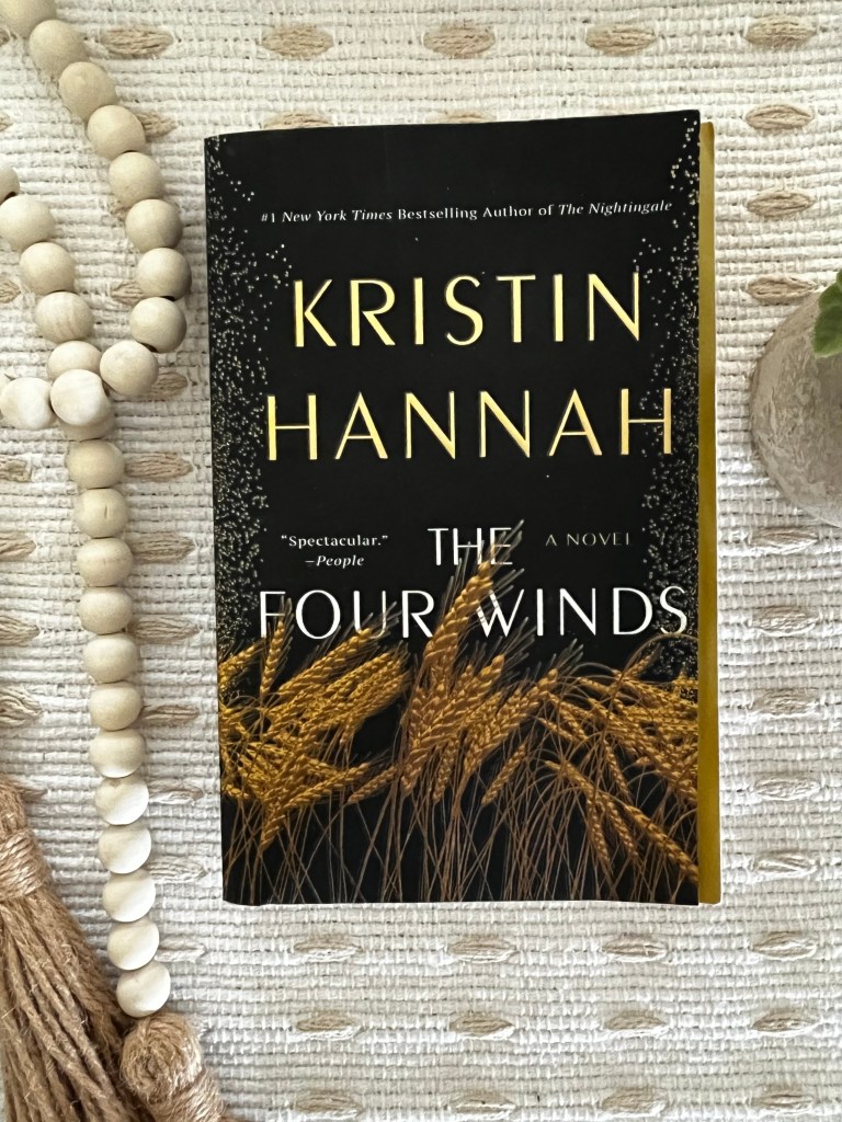 The Four Winds by Kristin Hannah displayed on a cream textured fabric with wooden beads and greenery.