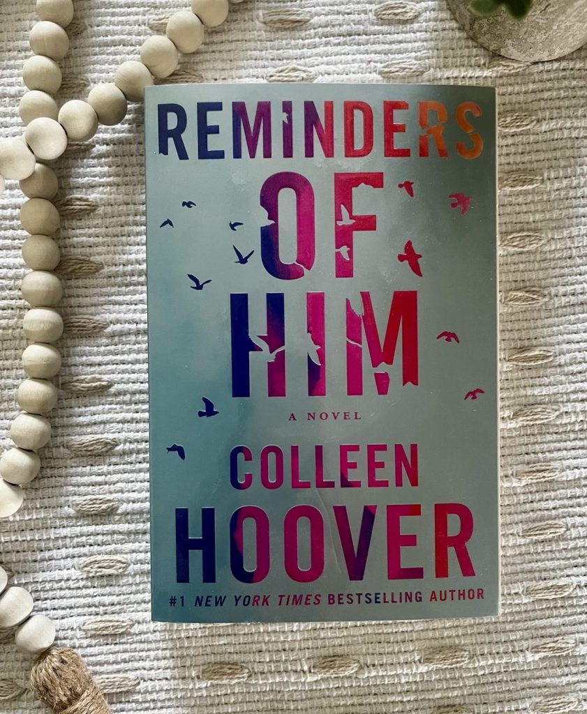 Reminders of Him by Colleen Hoover on a cream textured blanket with wooden beads Image Description: