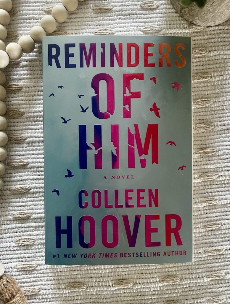 Reminders of Him by Colleen Hoover on a cream textured blanket with wooden beads Image Description: